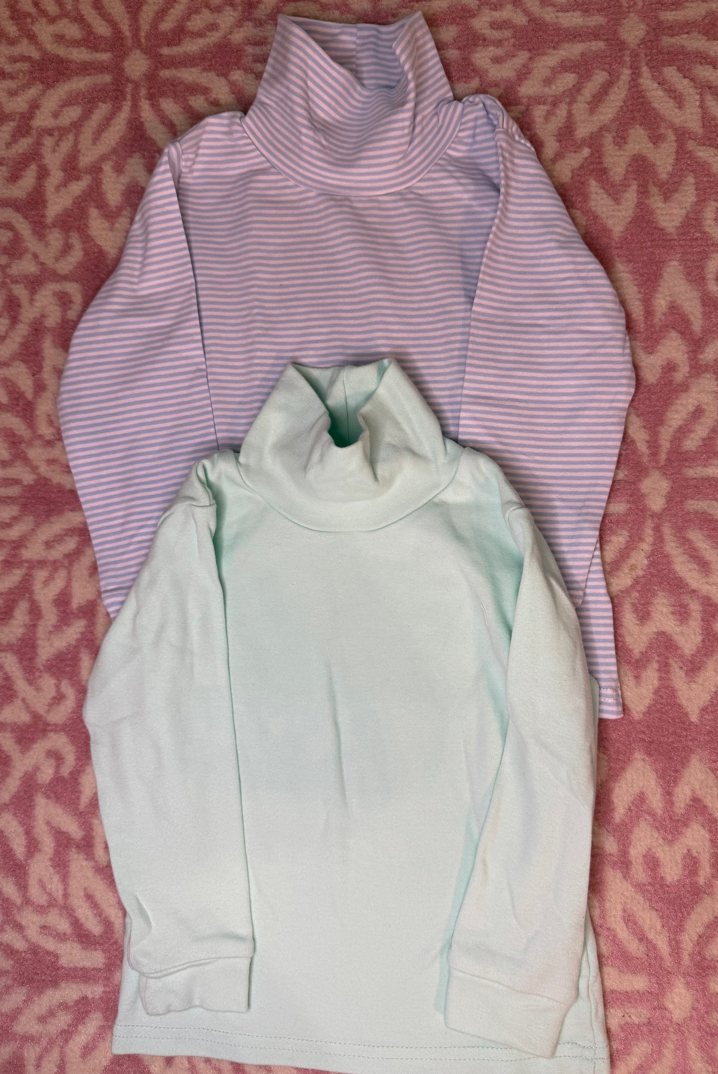 Two long sleeve children's turtleneck shirts, one mint green and one pink striped, on a pink patterned background.