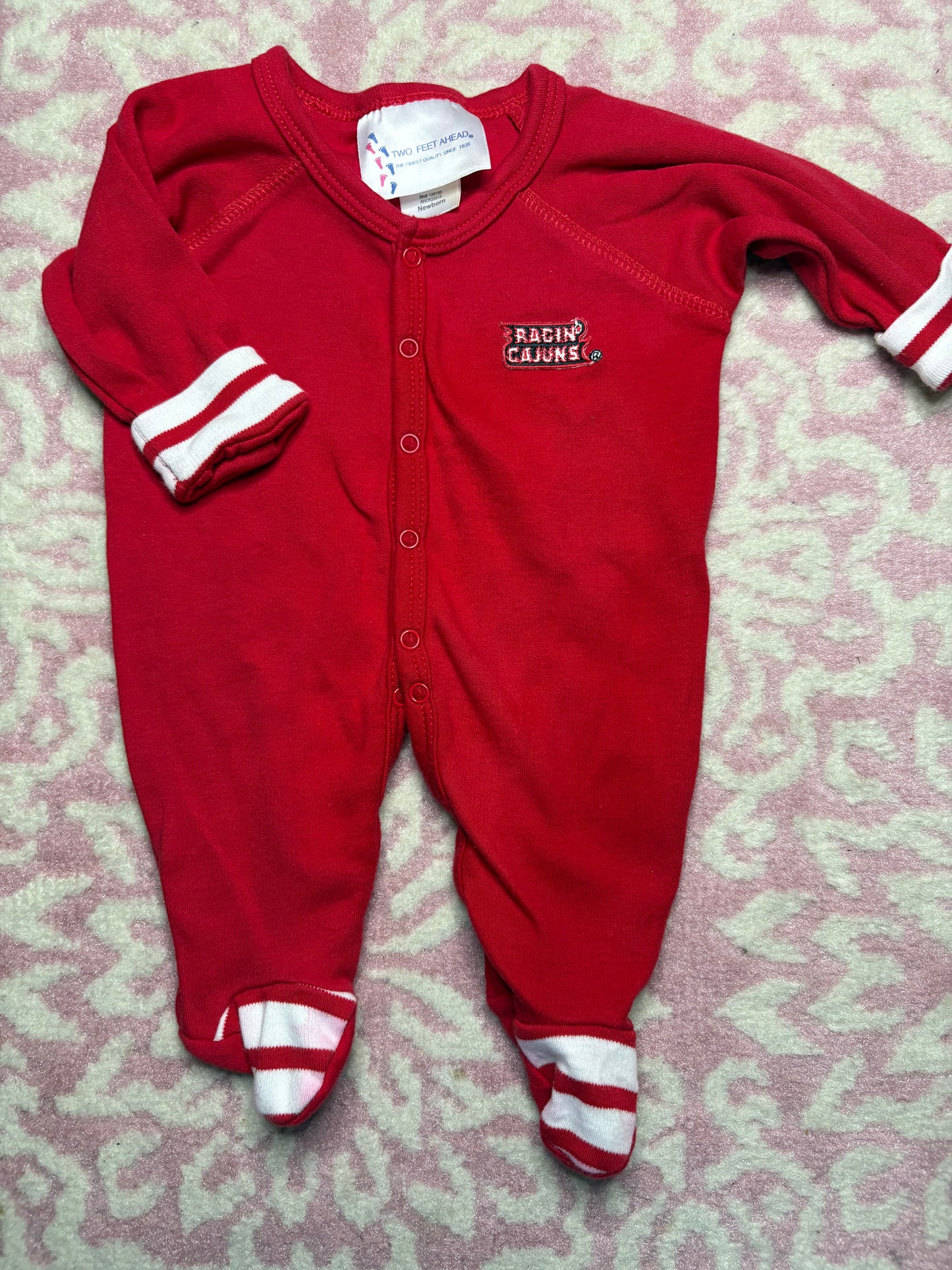 Red baby sleeper with white striped cuffs and feet, Ragin Cajuns logo on chest, laid on pink patterned blanket
