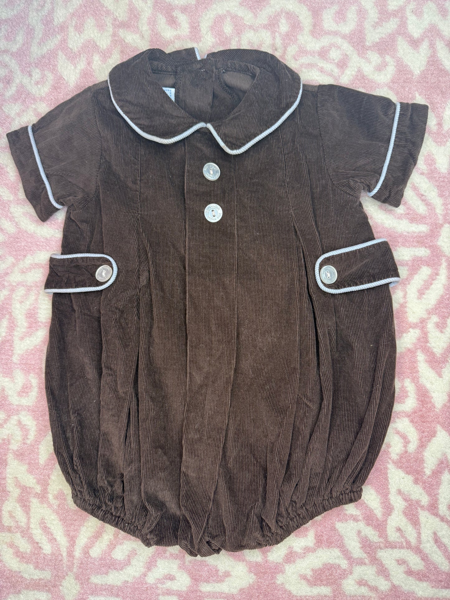 Brown corduroy baby romper with white trim and buttons on a pink patterned background