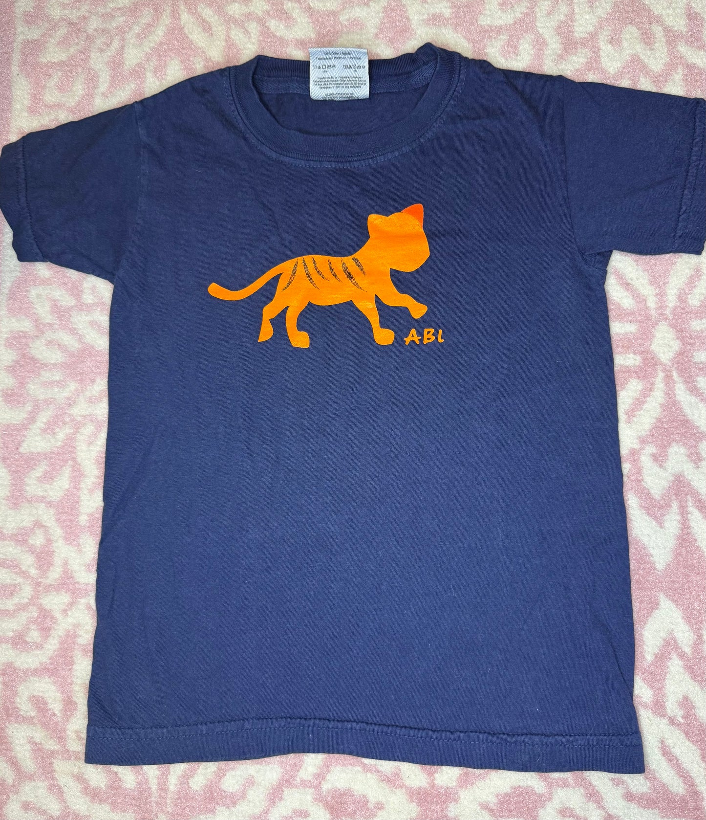 Navy blue children's t-shirt with orange tiger graphic and 'ABI' letters on pink patterned background.