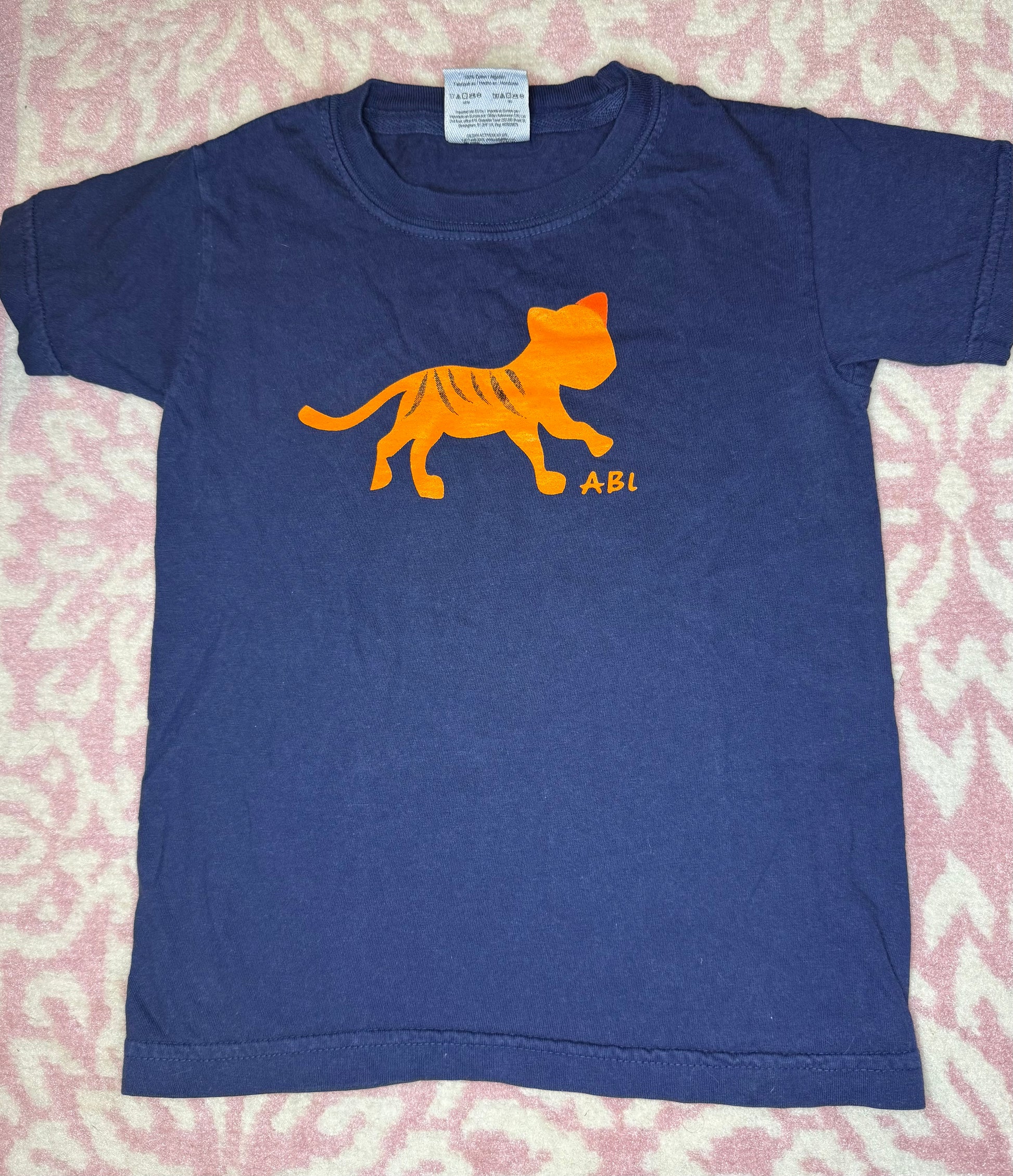 Navy blue children's t-shirt with orange tiger graphic and 'ABI' letters on pink patterned background.