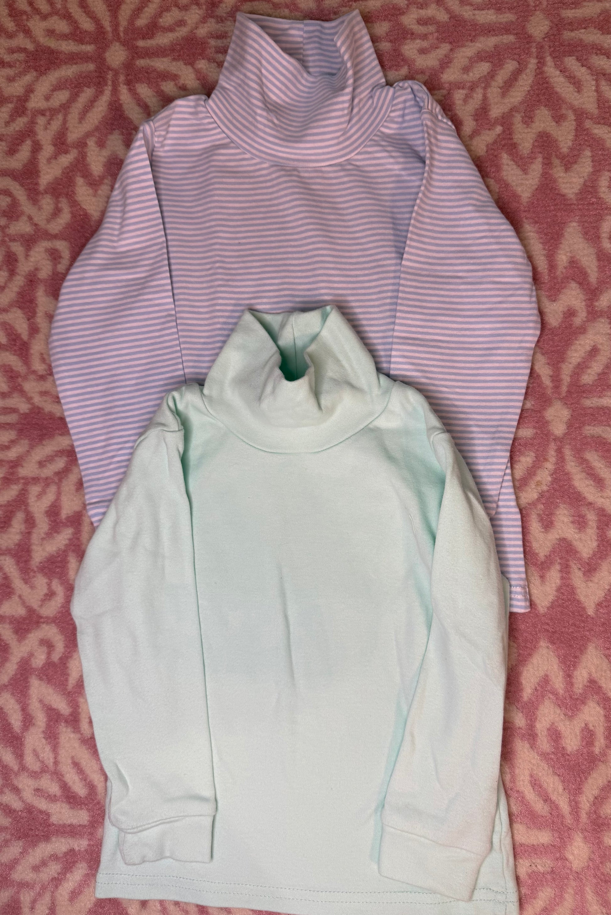 Two long sleeve children's turtleneck shirts, one mint green and one pink striped, on a pink patterned background.