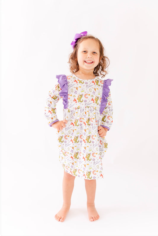 Smiling girl in long sleeve dinosaur print dress with purple ruffle accents and hair bow