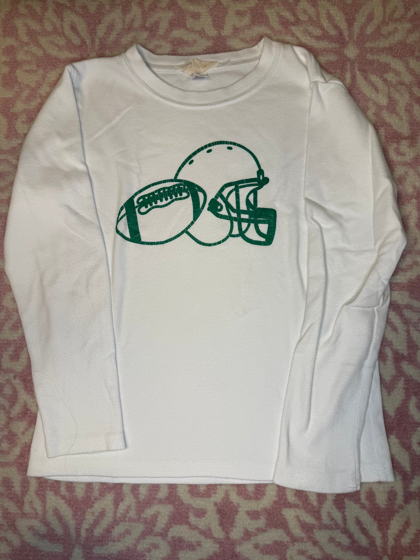 White kids’ long sleeve shirt with green football and helmet graphic, laid on pink patterned fabric.