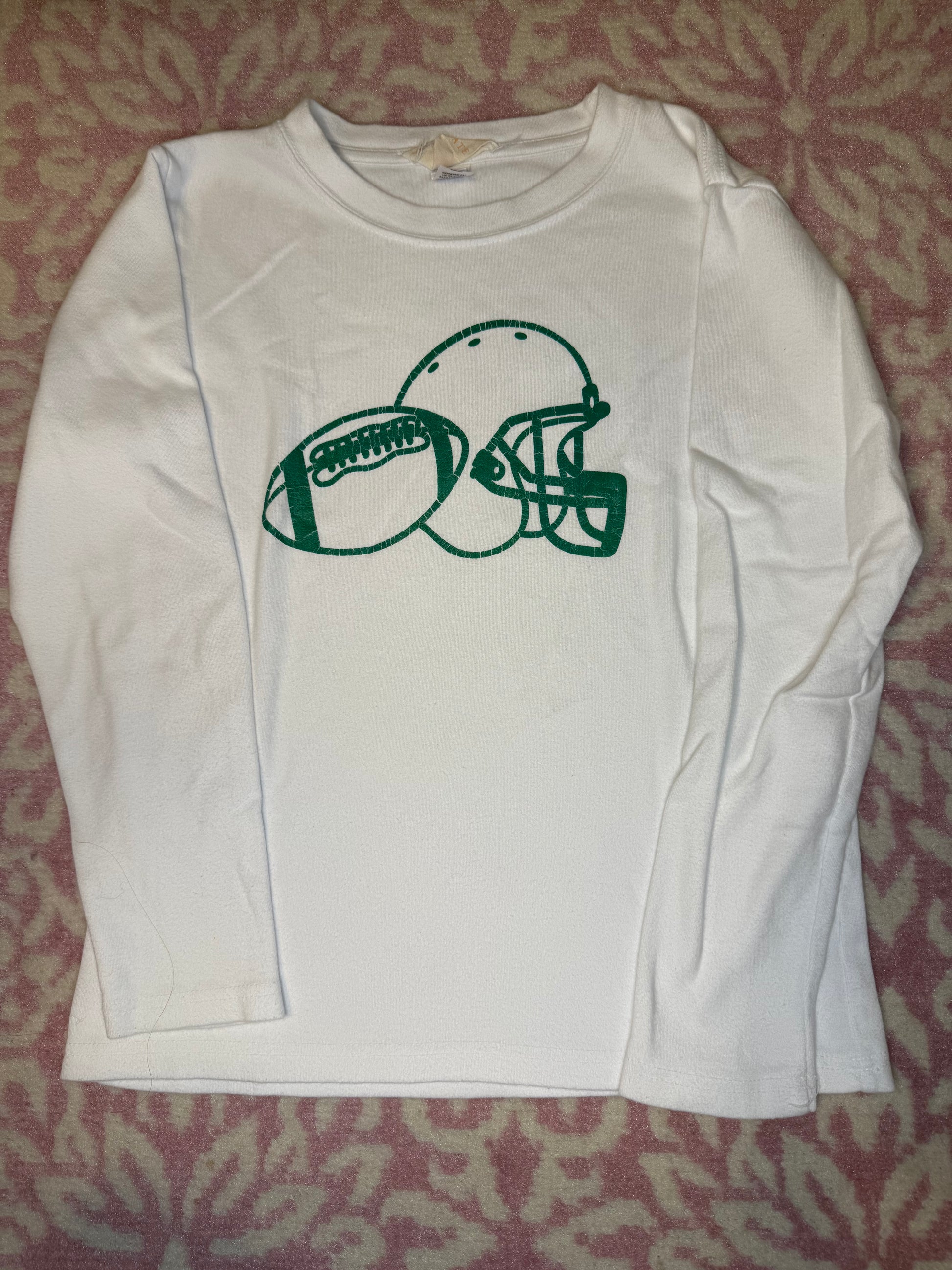 White kids’ long sleeve shirt with green football and helmet graphic, laid on pink patterned fabric.